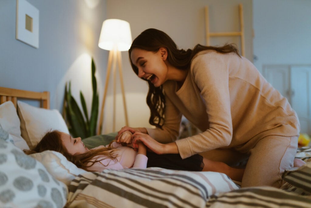 Cute mother in a beige sweater tickles and puts to bed a little daughter in the bedroom