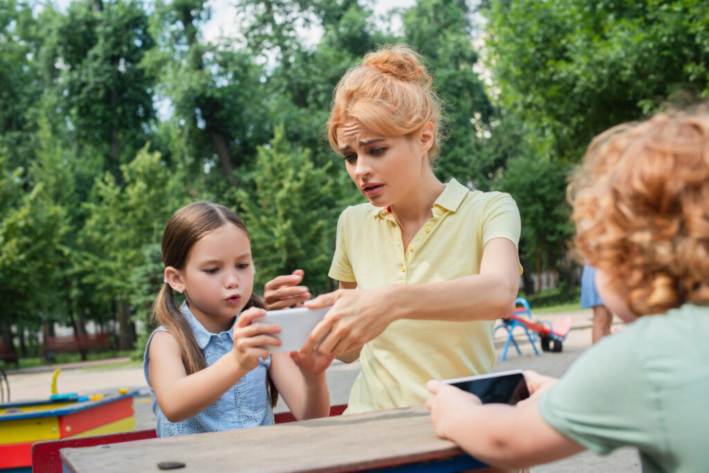 displeased woman taking smartphone from daughter while sitting on playground