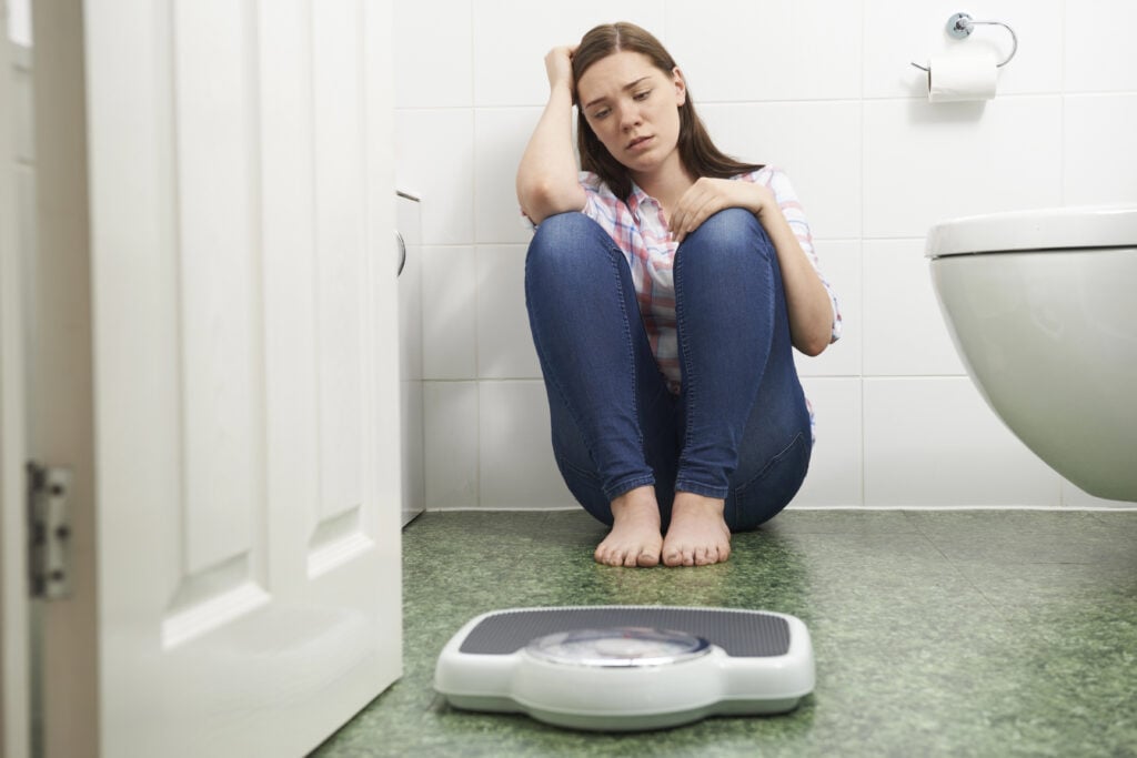 Unhappy Teenage Girl Sitting On Floor Looking At Bathroom Scales