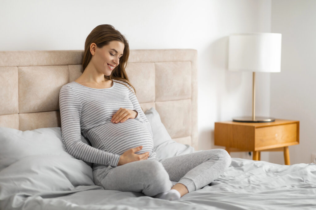 A serene pregnant woman sits comfortably on a bed, holding her belly 