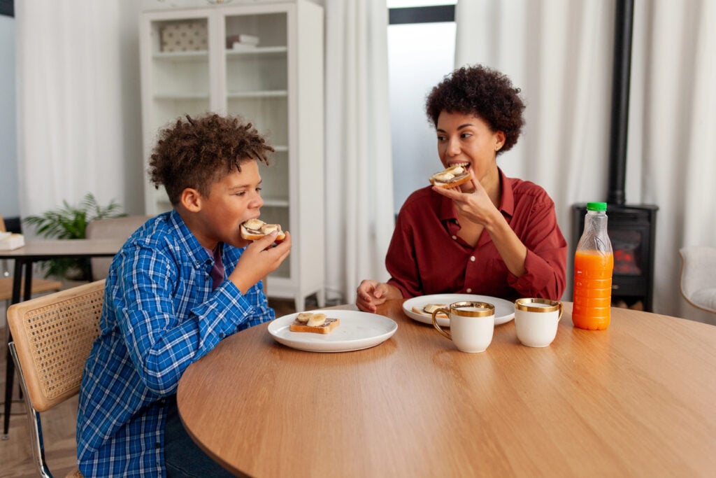 African American woman eating chocolate butter banana sandwiches and juice with her son at home at the table, teenager having lunch with his mother and laughing