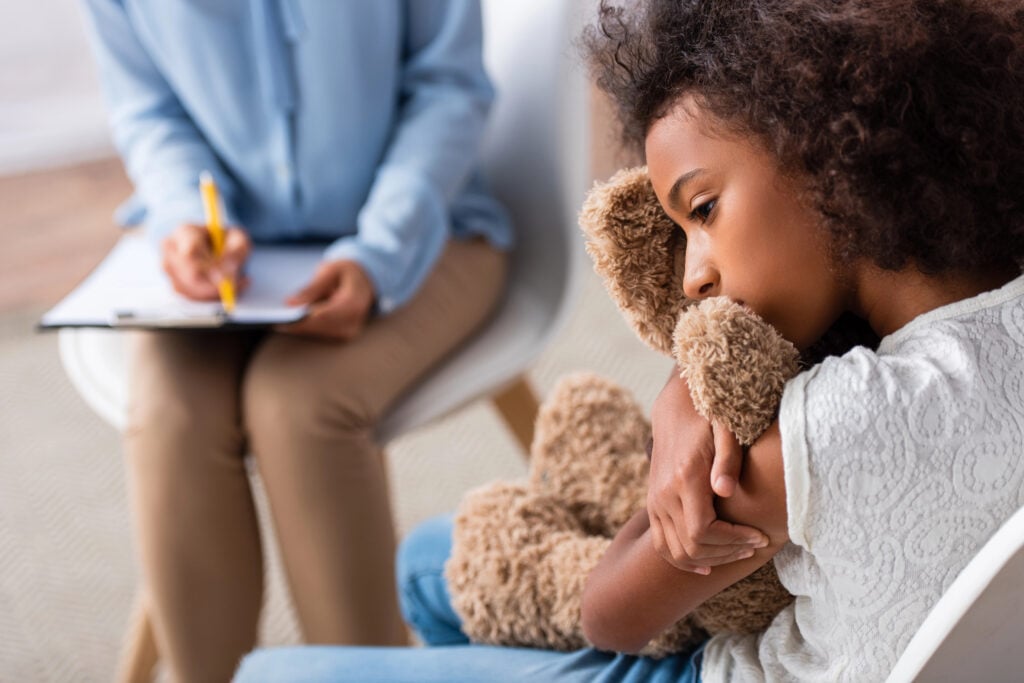 Upset african american girl with autism hugging teddy bear during consultation with blurred psychologist on background