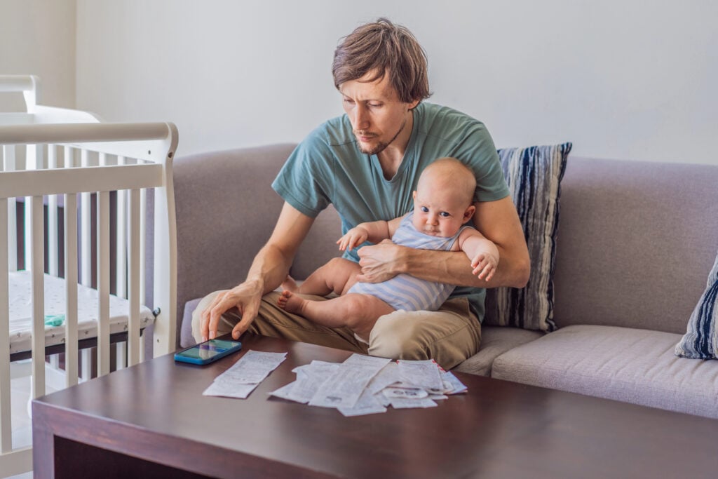 Father sitting on sofa holding baby while calculating monthly expenses with receipts at home. Stressed dad managing budget and childcare. Financial stress, parenthood and real life concept.