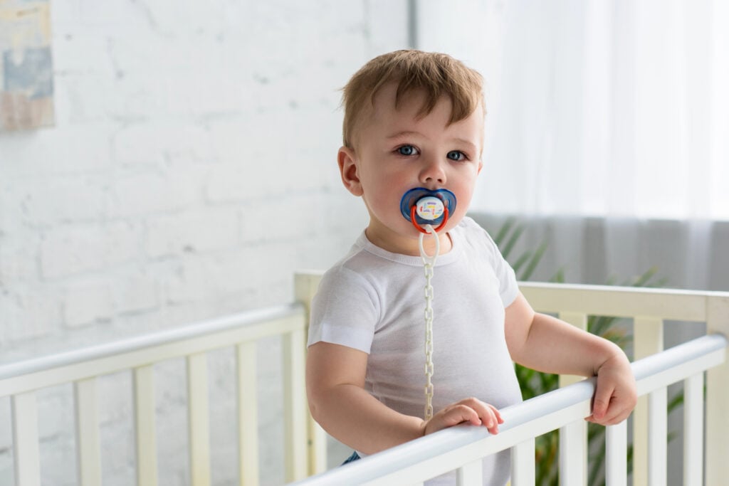 cute little baby boy with pacifier in baby crib looking at camera at home