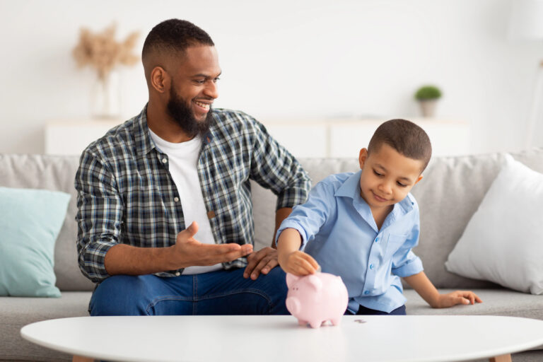 Black Boy And His Dad Putting Money In Piggybank Indoors