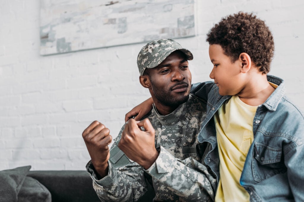 Father in army uniform and african american boy embracing and talking at home