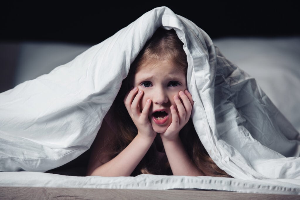 scared kid screaming and holding hands on face while hiding under blanket isolated on black