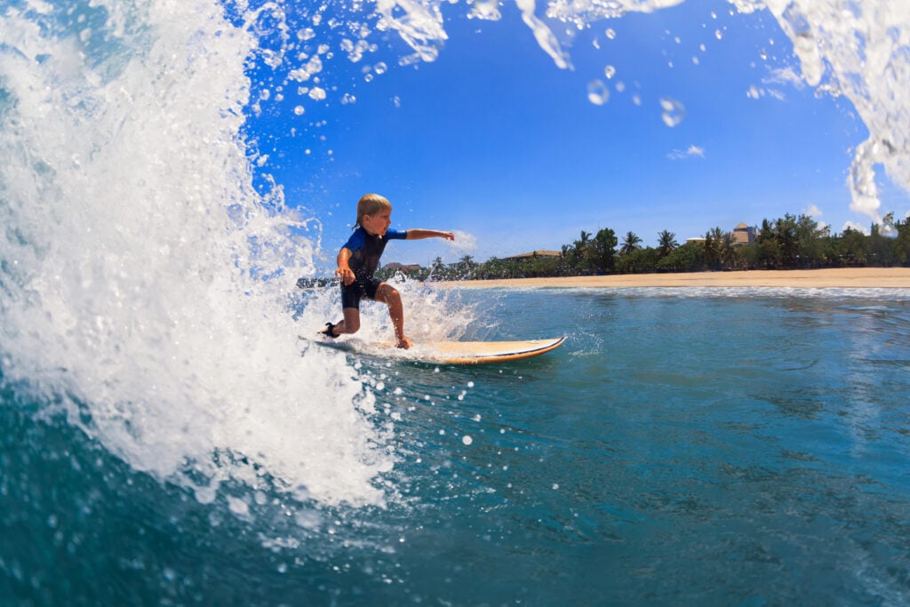 Happy surf boy - young surfer learn to ride on surfboard with fun on sea waves. Active family lifestyle, kids outdoor water sport lessons, swimming activity in surfing camp. Summer vacation with child