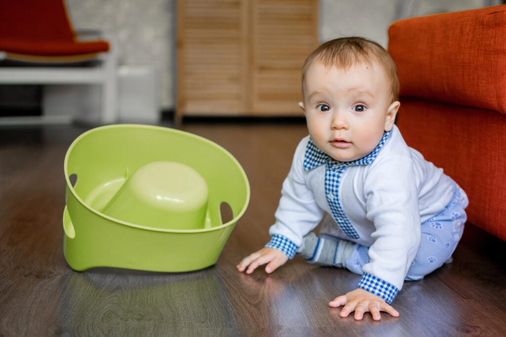 Portrait of an innocent baby boy sitting on floor in room near flipped over potty. "Oops". Little toddler with surprised face, toilet training gone wrong