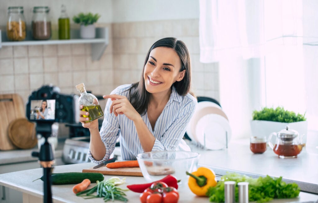 Professional beautiful happy young woman is blogging for her  kitchen channel about healthy living in the kitchen of her home and looking on camera on a tripod