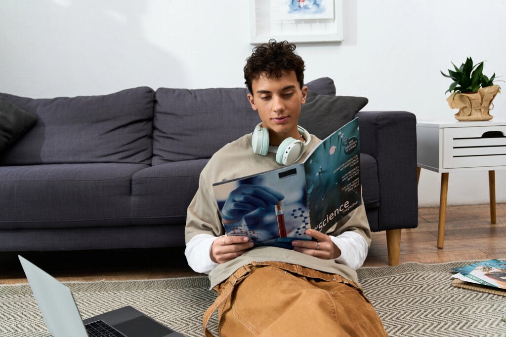 Teenage boy enjoying some leisure time while reading a magazine and using his laptop at home.