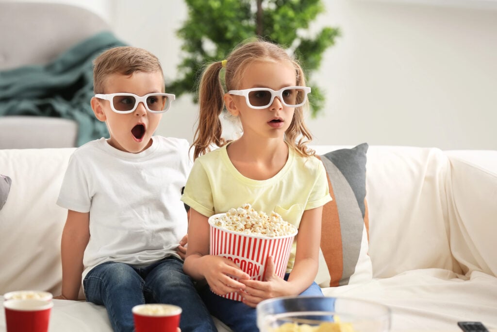 Cute children eating popcorn while watching TV at home