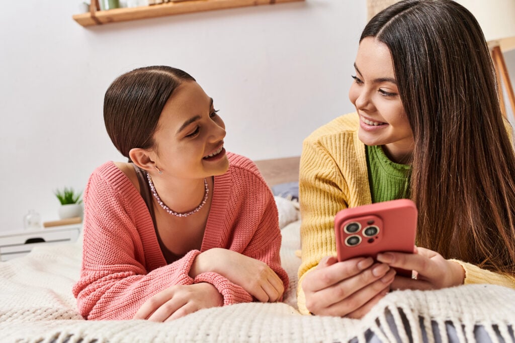 Two young women in casual clothing laying on a bed, engrossed in a cell phone, sharing a moment of connection and intimacy.