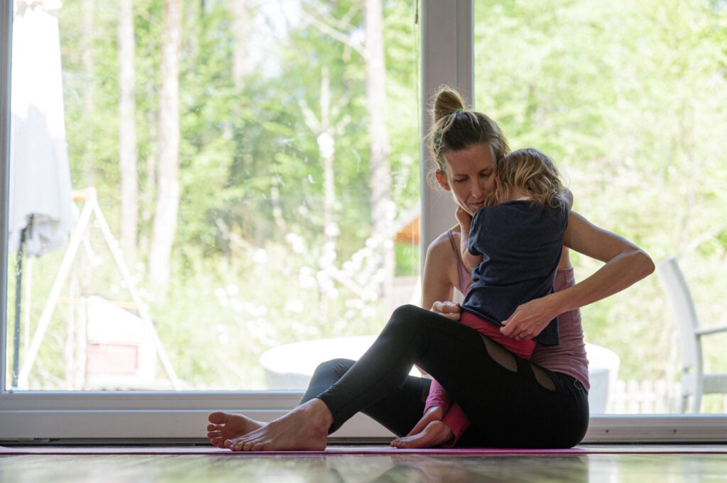 Young mother sitting on a fitness mat after home workout hugging her toddler daughter.
