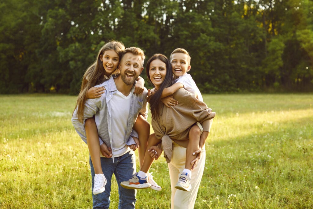 Portrait of happy smiling family of four with son and daughter standing on green grass in the summer park and looking cheerful at camera. Mother, father with two kids enjoying sunny day together.