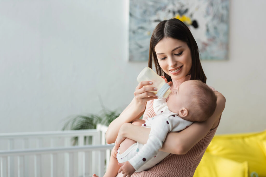 cheerful woman holding baby boy and feeding him with milk at home