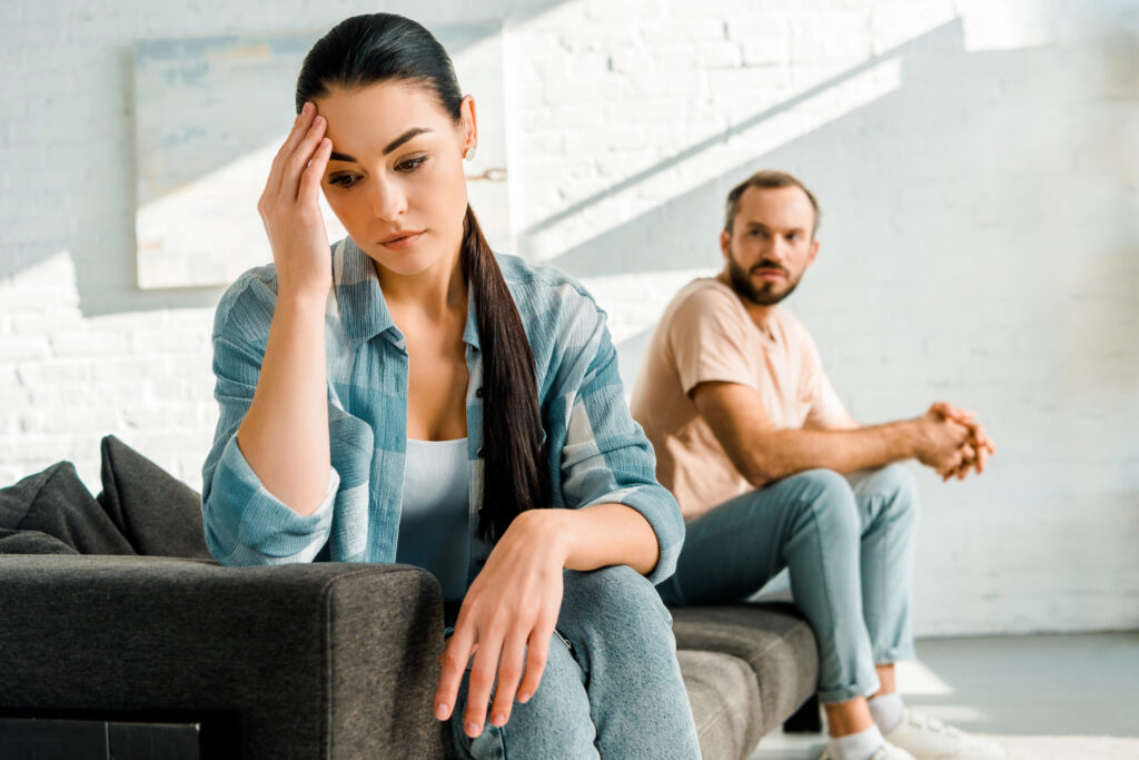 sad wife with hand on head sitting on foreground after arguing with husband at home