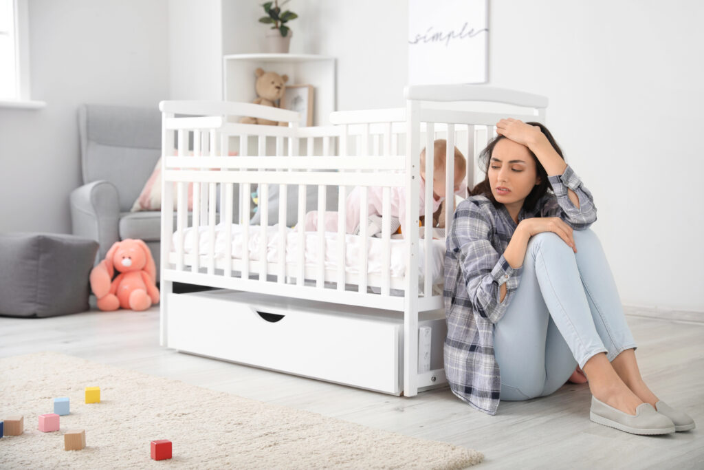 Young woman suffering from postnatal depression near bed with baby at home