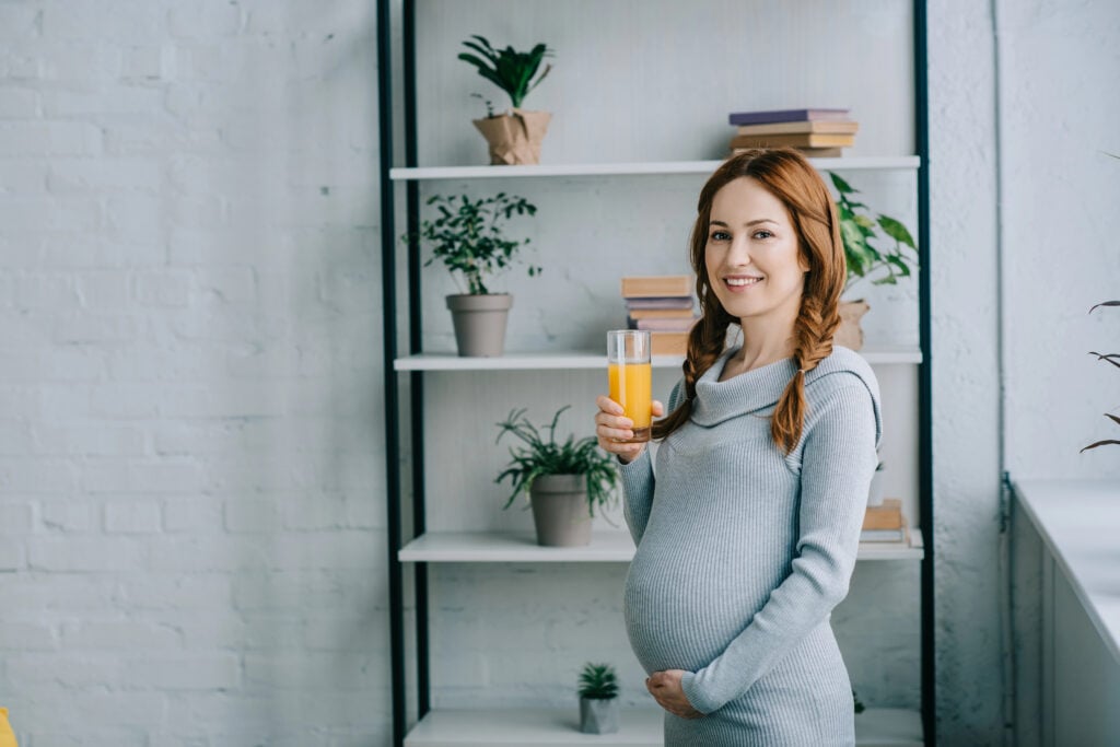 attractive smiling pregnant woman holding glass of orange juice and looking at camera at home