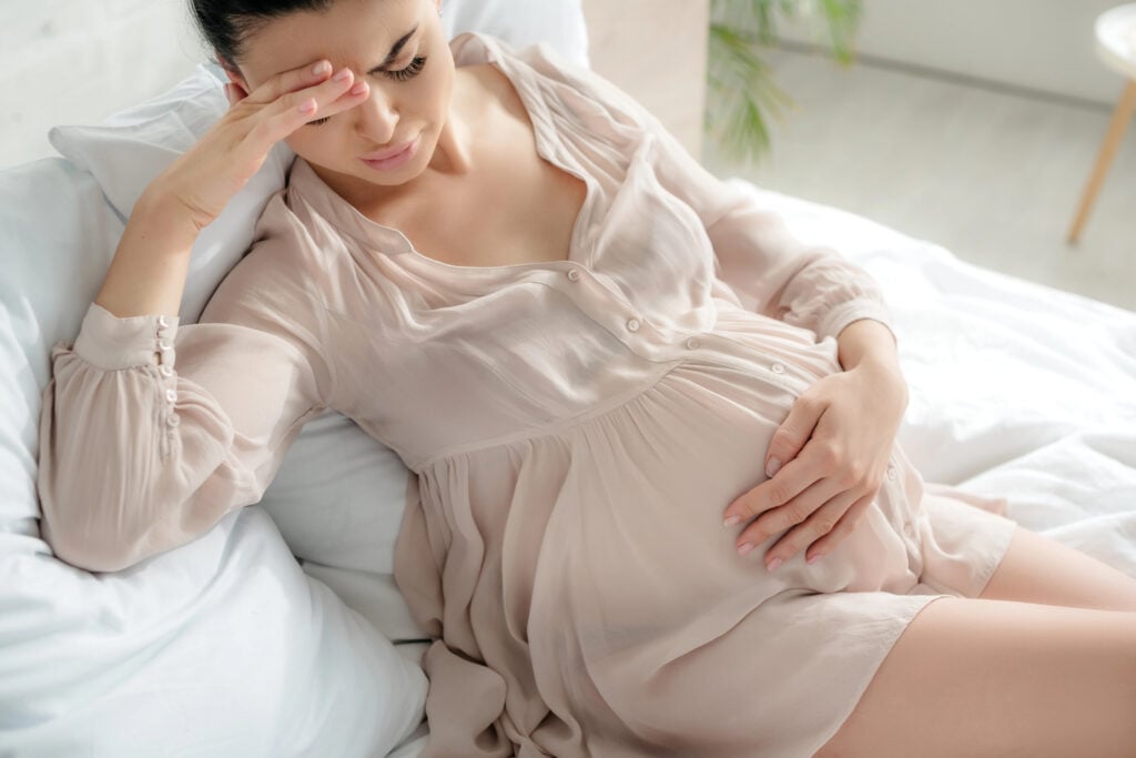 exhausted pregnant woman in nightie having headache and touching tummy while sitting on bed