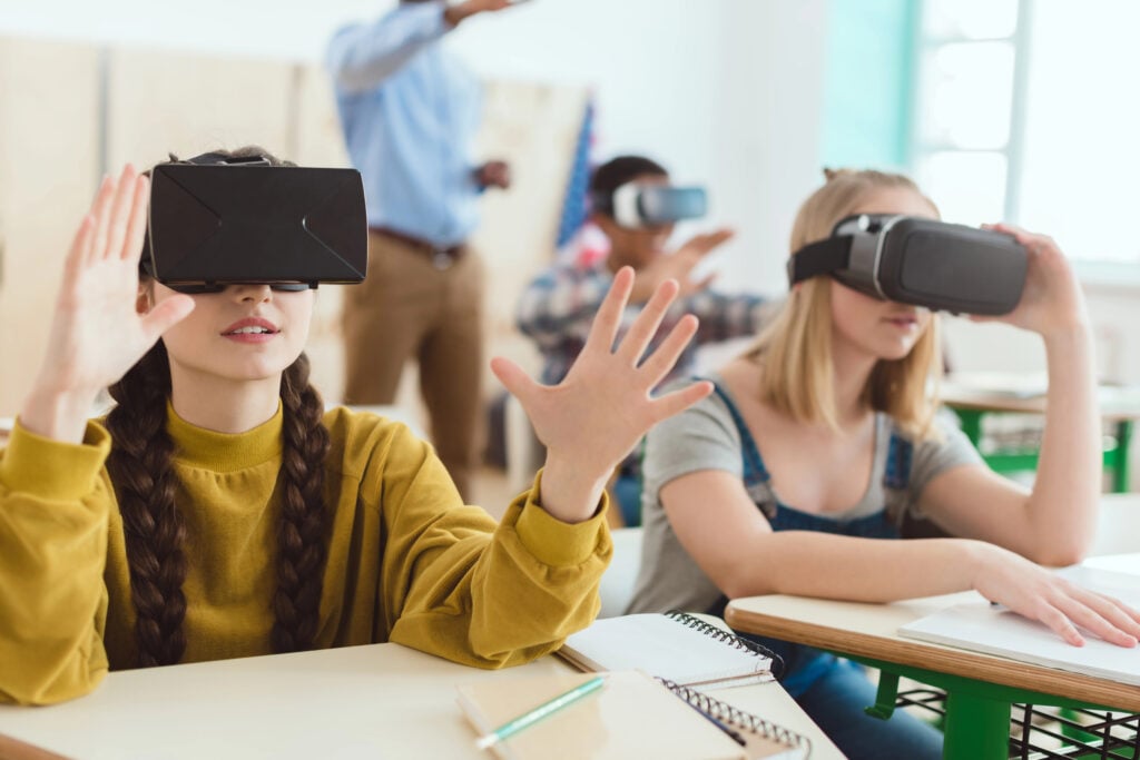 Two teenage schoolgirls using virtual reality headsets and classmate with teacher behind