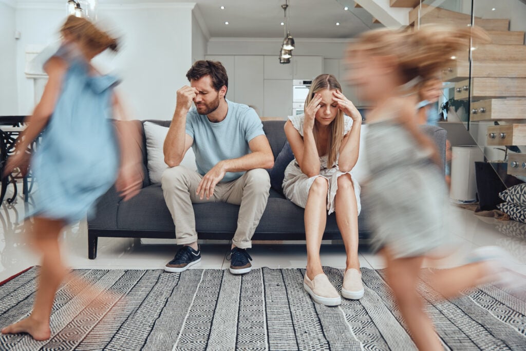 Kids running, parents on sofa tired and chaos in living room with frustrated mom and dad burnout. Happy children, parenting stress and excited girl and boy with energy play, woman and man in crisis.
