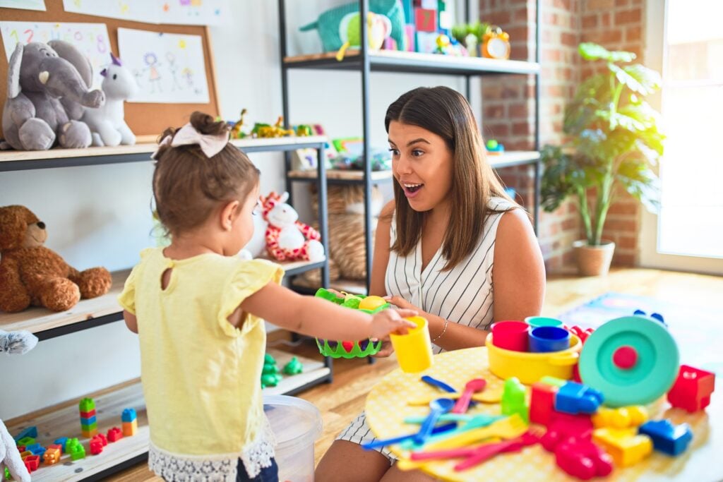 Young beautiful teacher and toddler playing with dishes, cutlery and cups toy on the table at kindergarten