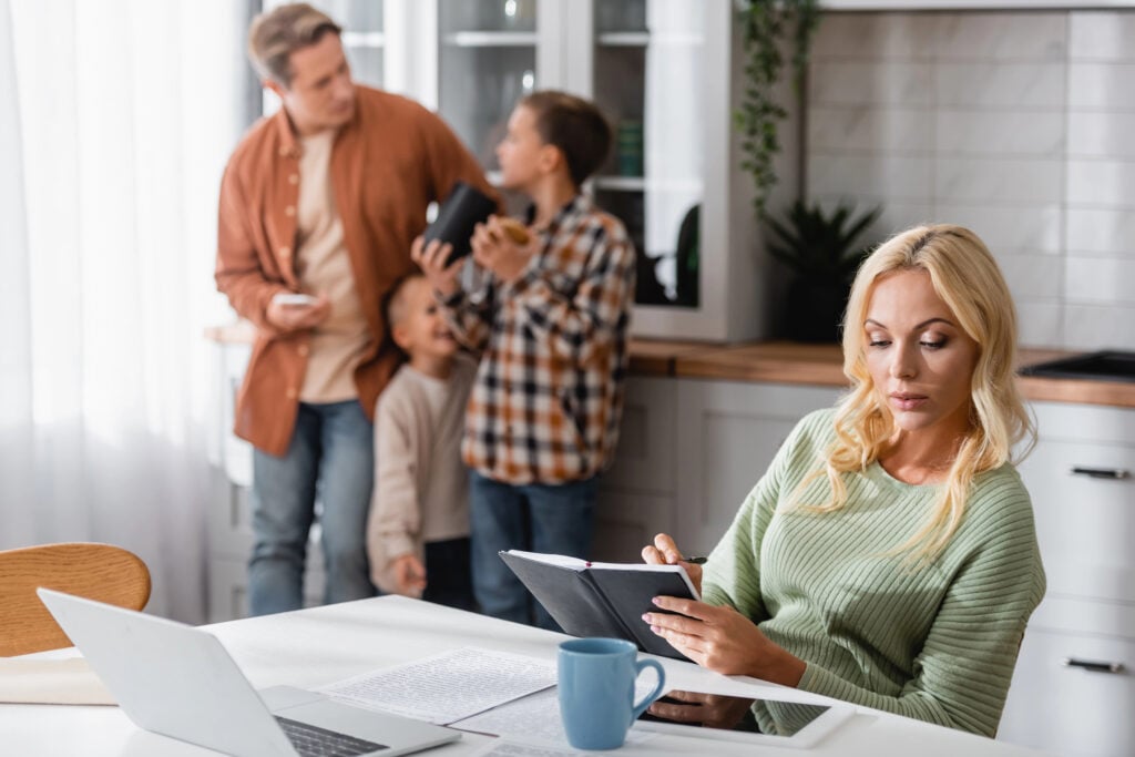 woman writing in notebook while working in kitchen near husband and sons on blurred background