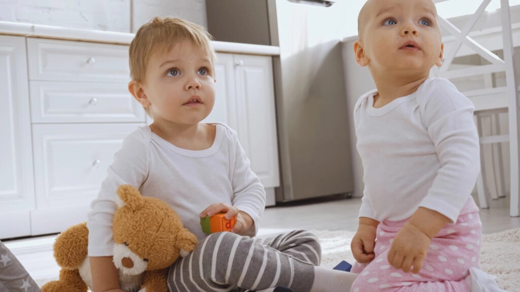toddler boy sitting on carpet with teddy bear near baby sister and looking away