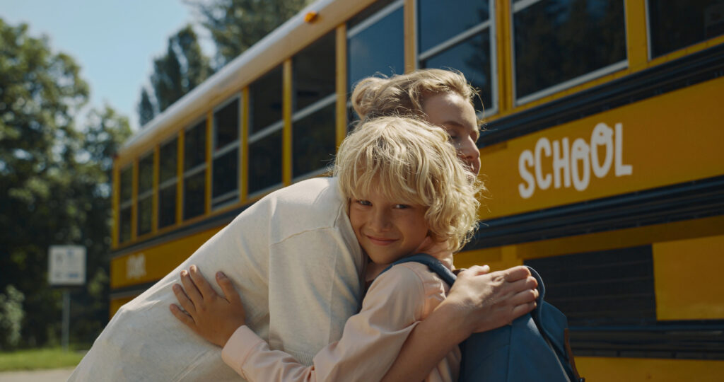 Young smiling mom hugging little son standing near yellow school bus. Joyful curly boy with backpack saying goodbye to cheerful mother before boarding sunny morning. Happy woman waving to schoolboy.