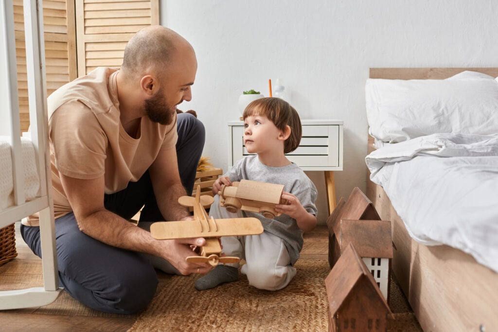 joyful modern father and son in cozy homewear playing with wooden toys and looking at each other