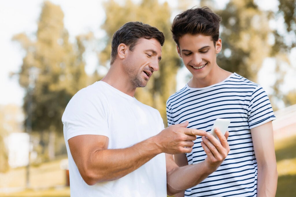 man pointing with finger at smartphone near teenager son in park