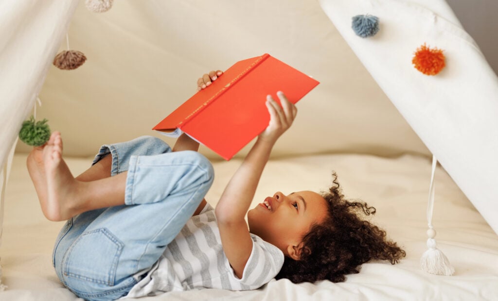 Portrait of cute little african american kid curly boy with book smiling at camera while   reading book  in play tent at home, happy child playing alone in children room