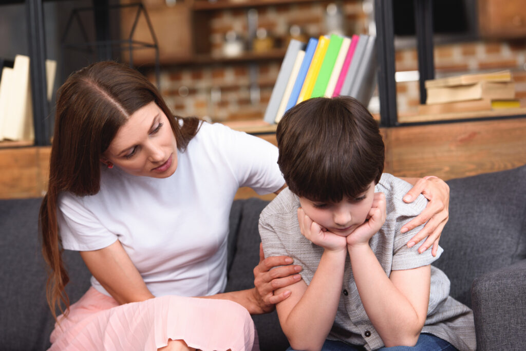 selective focus of mother cheering up depressed little son on sofa at home