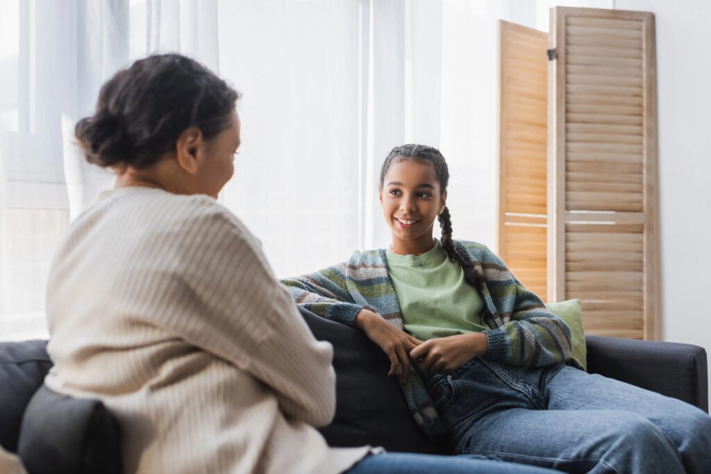 smiling african american teen girl talking with blurred mom on sofa at home