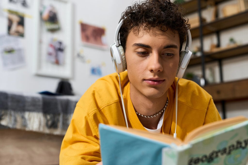 A trendy teenager relaxes at home, reading a book while listening to music through headphones.