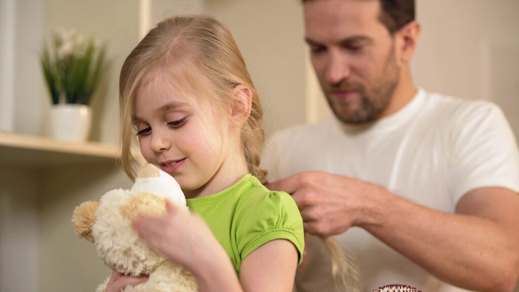 Happy daughter waiting father braiding her blonde hair, playing with teddy-bear