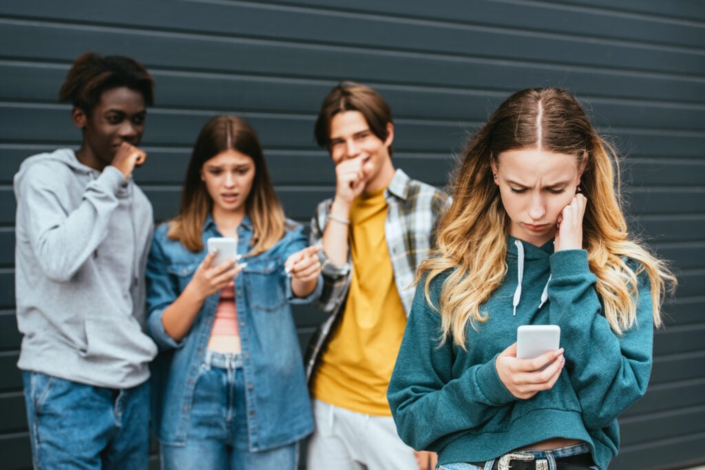 Selective focus of upset girl using smartphone near laughing multiethnic teenagers outdoors