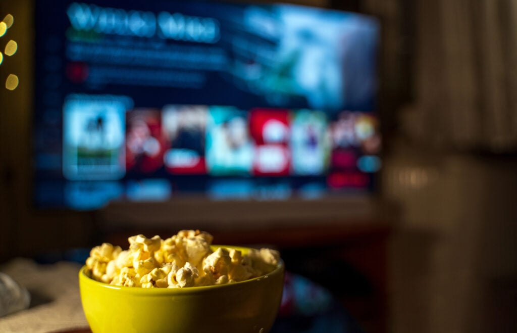 A bowl of popcorn in the foreground with a TV in the background showing a streaming service interface.