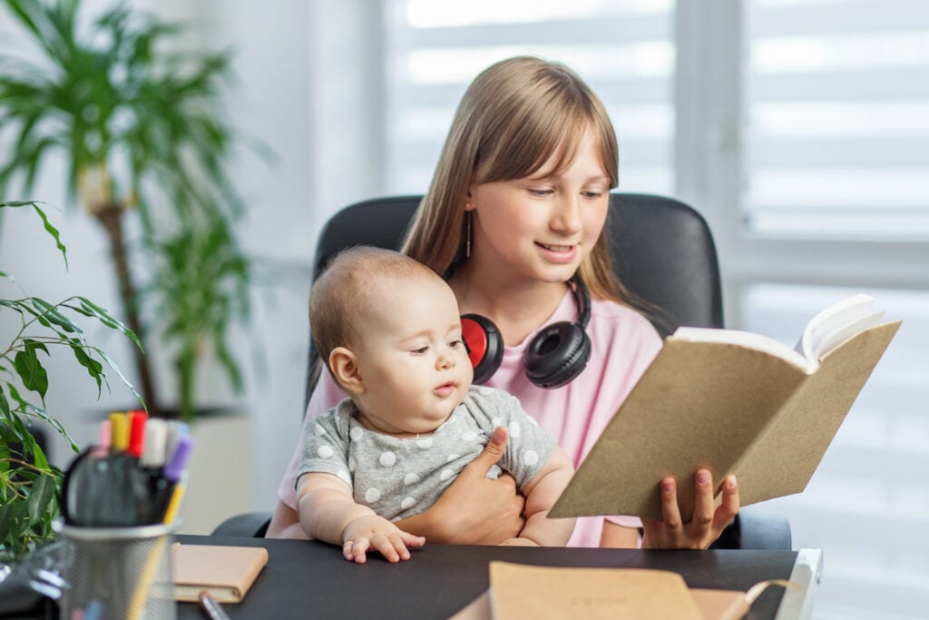 Smiling girl holds baby on her lap while reading a book aloud at home, showing family bonding and early childhood learning.
