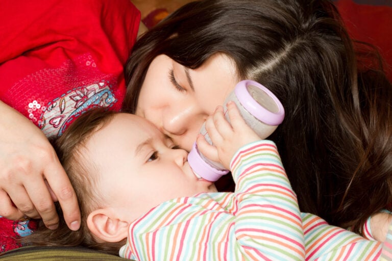 Young mother feeds child girl with a bottle with infant formula on bad. The