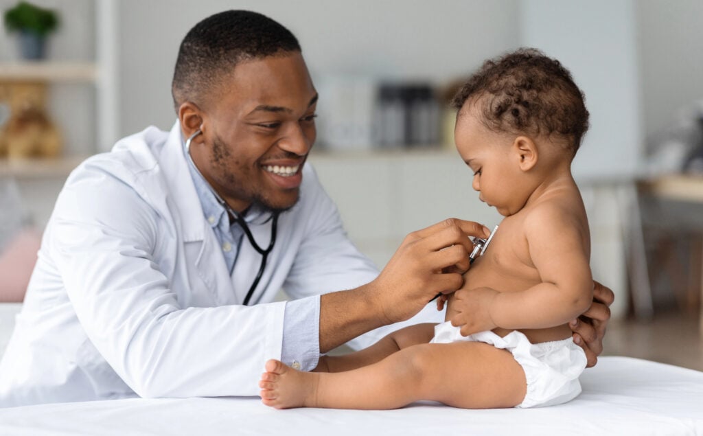 Black Pediatrician Doctor Doing Check Up To Cute Newborn Baby In Diaper, African American Pediatrist Using Stethoscope For Listening Childs Heartbeat, Checking Infants Heart And Lungs In Clinic