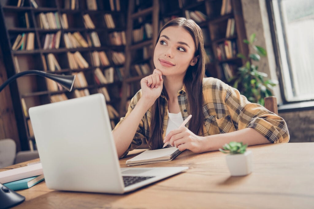 Portrait of attractive dreamy cheery girl learner doing home task using laptop fantasizing at loft industrial interior indoors