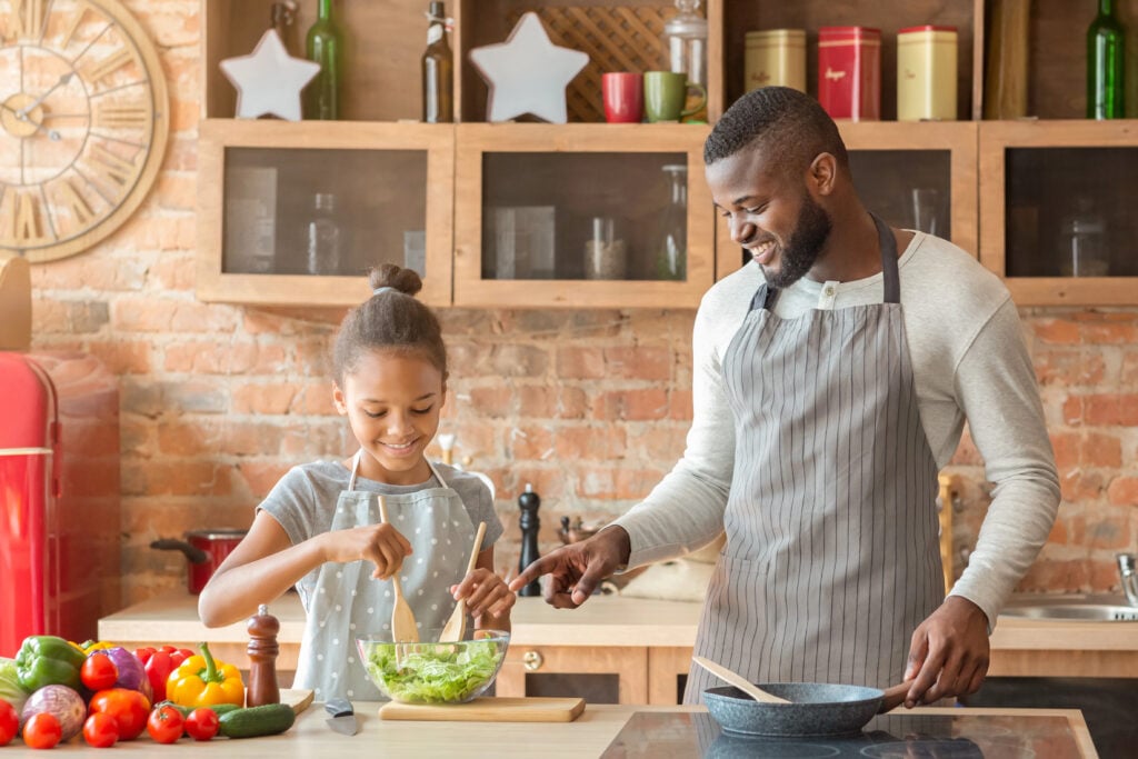 Cute girl mixing healthy veggies salad, father teaching her