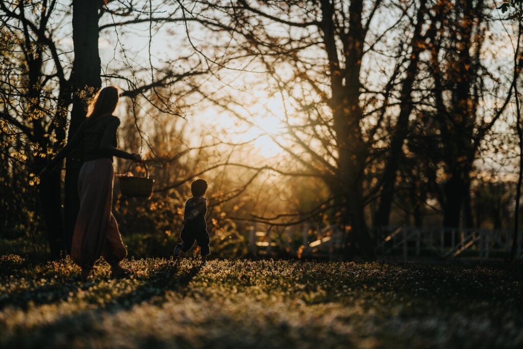 A mom walks with a child across a sunlit park meadow at sunset. Golden hour light silhouettes the mother and child among trees and grass.