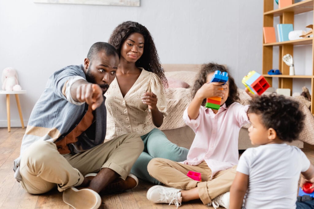 african american man pointing with finger near wife and children playing with building blocks on floor