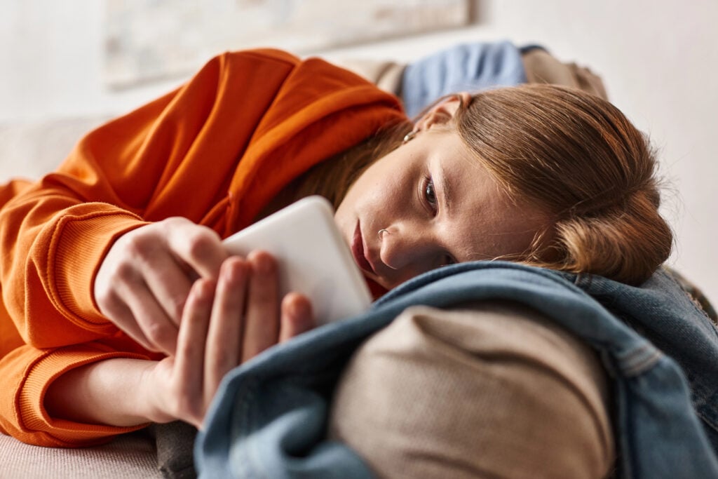 troubled teenage girl using her smartphone and feeling down while lying on sofa in living room