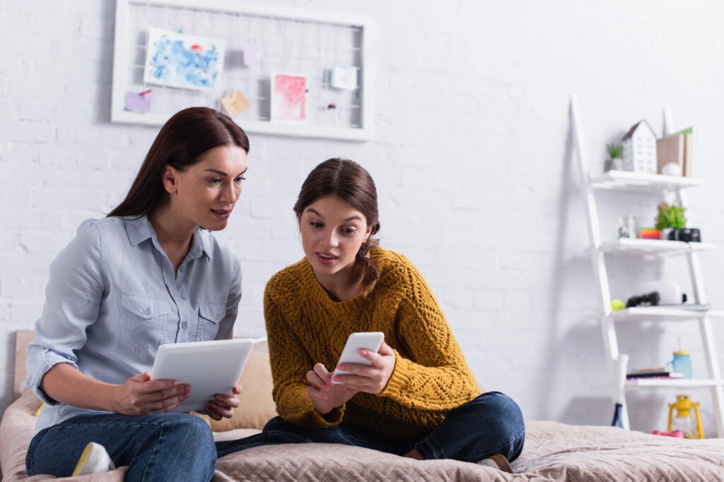 teenage girl holding smartphone near mother with digital tablet on bed