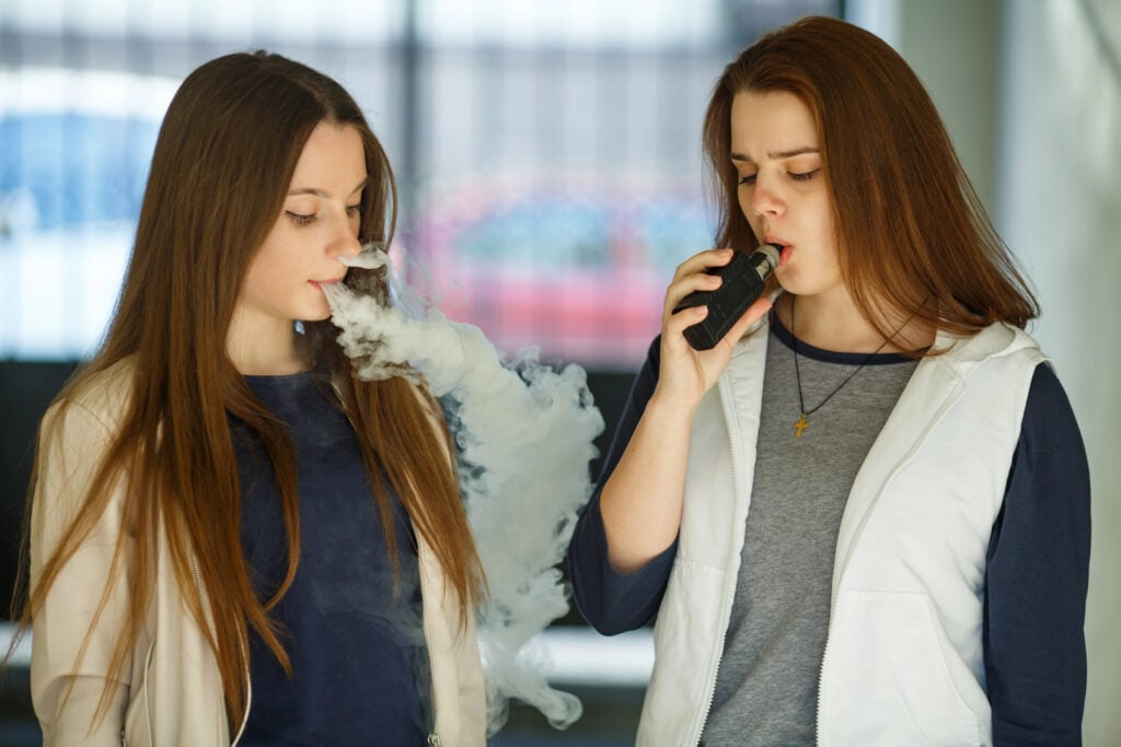Vape teenagers. Two young cute girls in casual clothes smoke electronic cigarettes outdoors in the street in summer day. Bad habit that is harmful to health. Vaping activity.