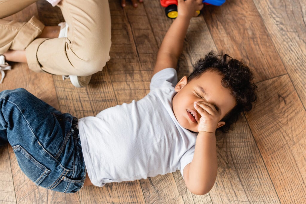 top view of sleepy african american boy touching eyes while lying on floor at home